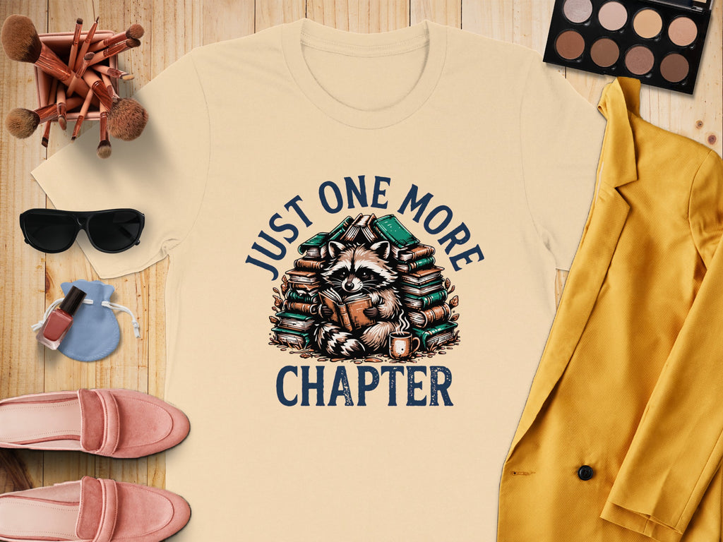 Light beige t-shirt with raccoon reading books graphic that says "Just One More Chapter".