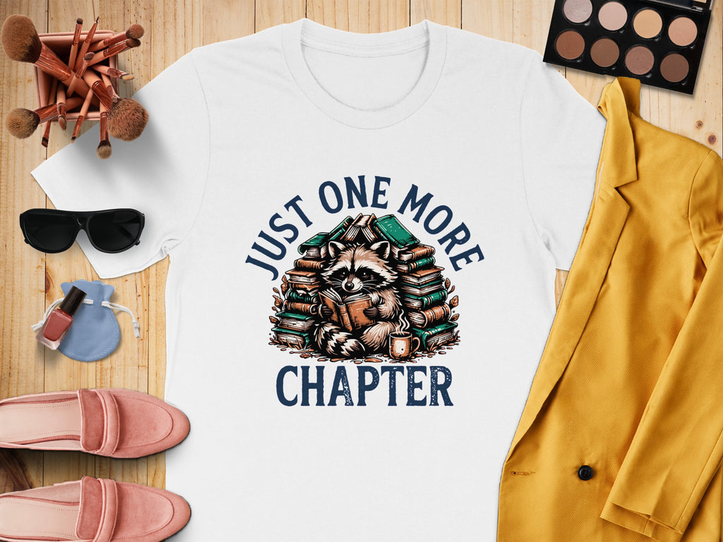 White t-shirt with raccoon reading surrounded by books, text "JUST ONE MORE CHAPTER".