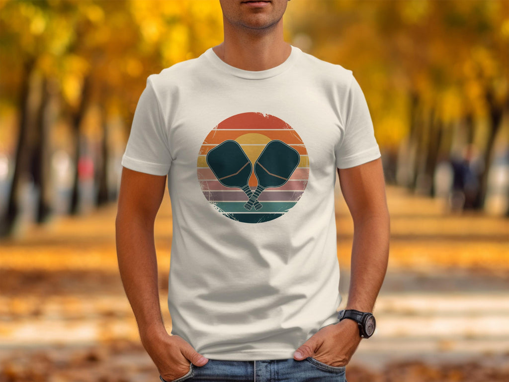 White t-shirt featuring crossed pickleball paddles on a distressed circular sunset graphic.