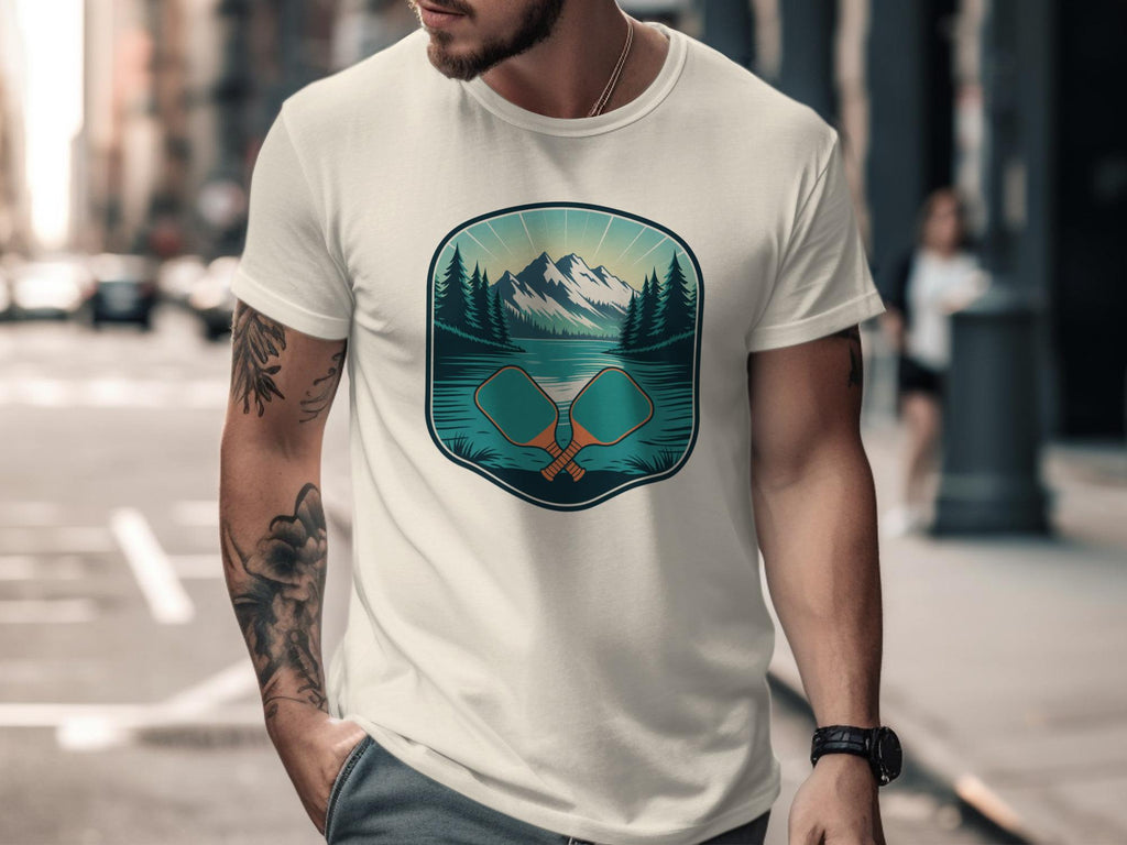 Cream t-shirt with a graphic of crossed pickleball paddles in a mountain landscape.