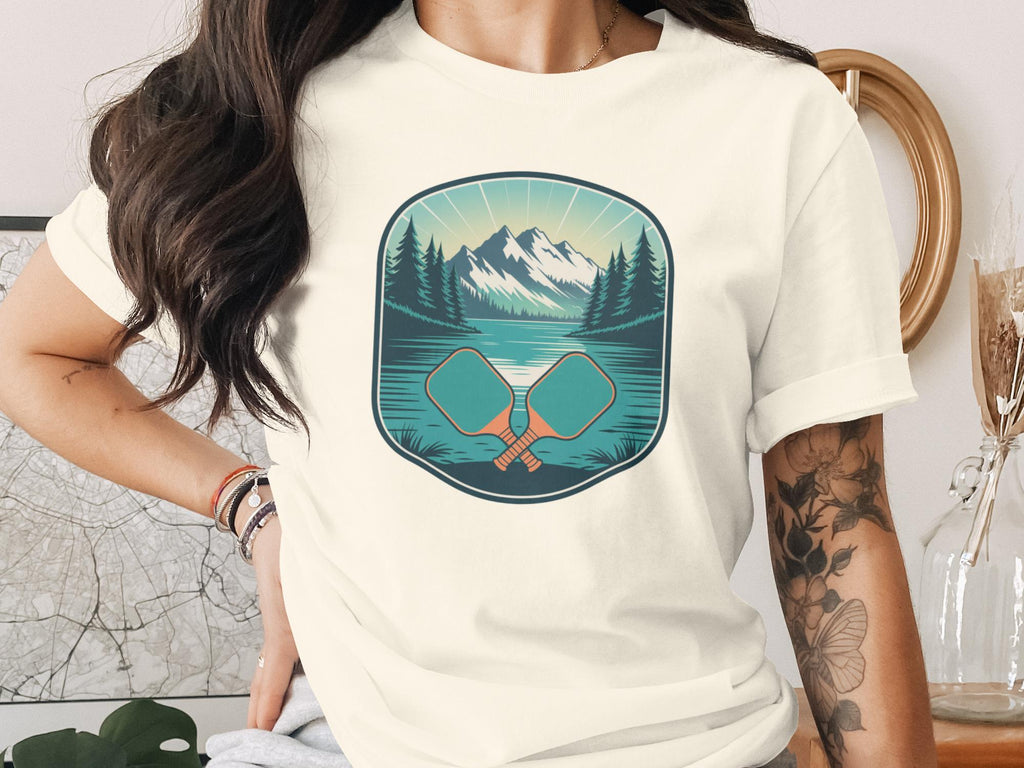 T-shirt graphic with two crossed pickleball paddles over a lake and mountains.